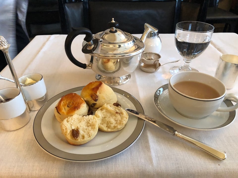 A white tablecloth table set for afternoon tea. In the center, a silver teapot with a curved black handle is surrounded by a sugar bowl, cream jug, and tea strainer. On the left, a small dish of butter sits beside jam. A plate in the foreground holds three golden-brown raisin scones, one split open. To the right, a teacup filled with milky tea rests on a saucer beside a silver-handled knife. A glass of water and other silverware complete the elegant arrangement.
