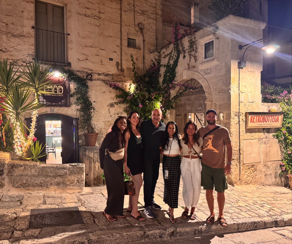 Nadia stands on the left with a group of five other people on a picturesque stone street at night, surrounded by historic buildings, greenery, flowers, and warmly lit restaurants.