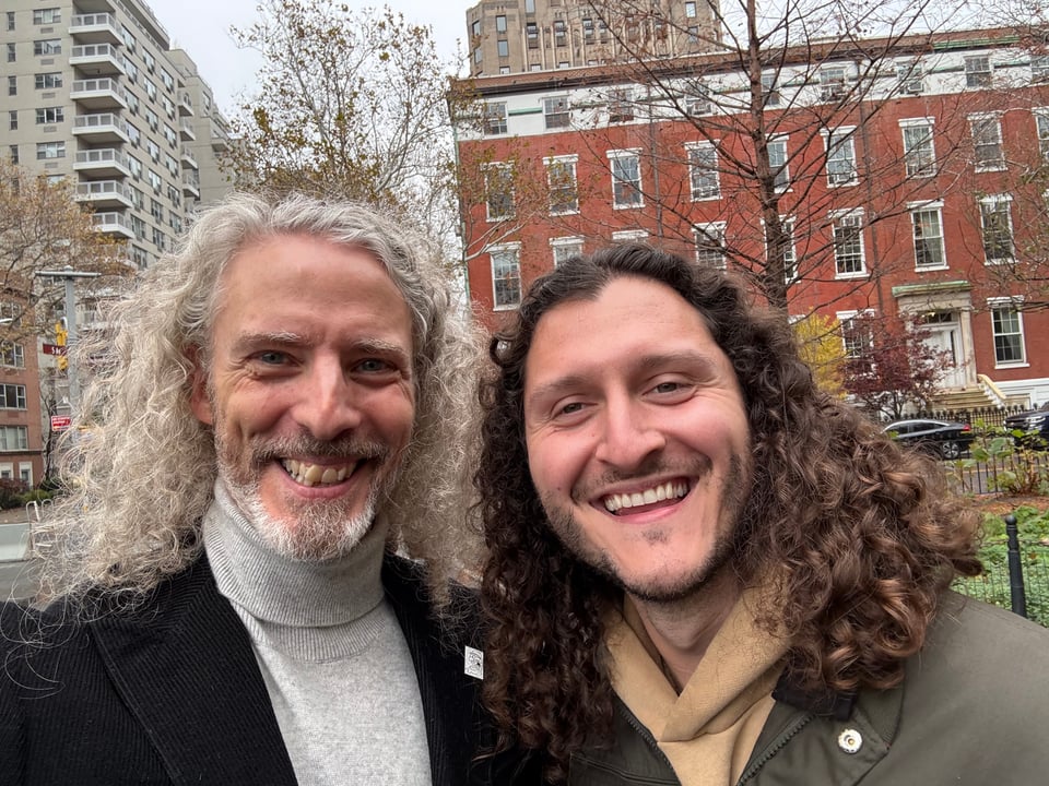 double-selfie of two white men with long hair, smiling effusively. They're in Washington Square Park in NYC, and there's an overcast sky behind them.