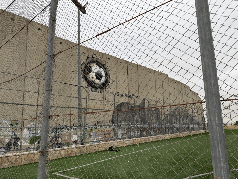 A close-up of the wall seen next to the soccer field in the previous photo. It includes a painting of a person who is lying down, with the words 'Save Aida Pitch'. It also includes a painting of a soccer ball that looks like it has crashed into the wall and smashed it.