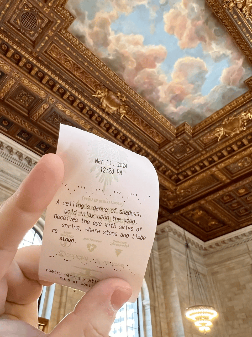 A picture of a hand holding up a piece of cash-Register-STYLE thermal paper. In the background, you see the ceiling of an old building with gold gilt around the edges and a center painted to look like a blue sky with clouds. On the piece of paper there is a date, March 11, 2024, 12:28 PM. A poem is printed that reads: "a ceiling's dance of shadows / Gold inlay upon the wood / Deceives the eye with skies of spring, / Where stone and timbers stood.'"