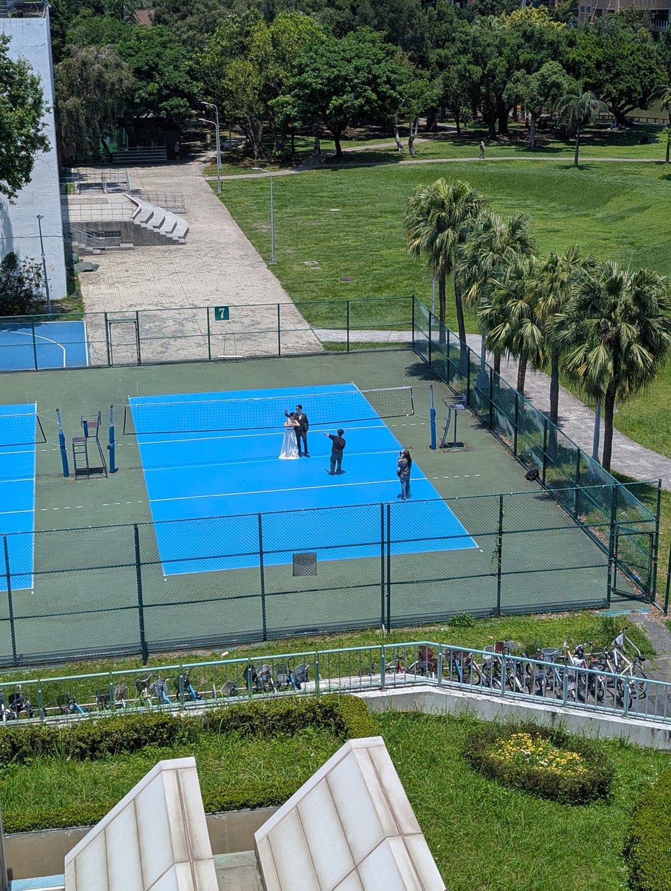 A bright blue volleyball court seen from an elevate distance. On it, a couple in a wedding dress and tuxedo are posing behind the net while a person directs them. A fourth person is taking photos.