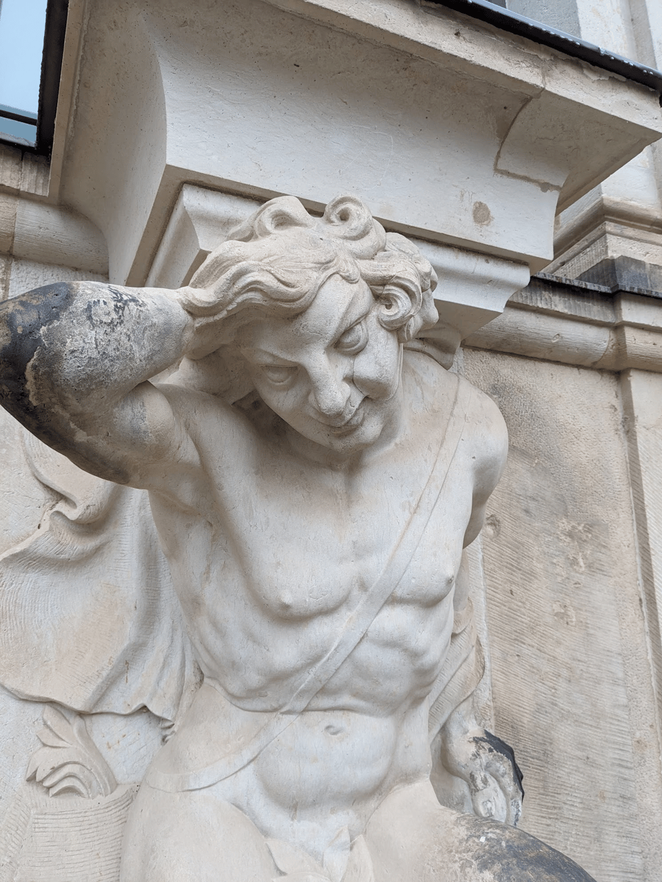 A male torso sculpted in the classical style. The figure is nude except for a fig leaf. He has short, curly hair and a devious expression. His right hand is on the back of his head, and his elbow is toward the viewer. The elbow is sooty, while the rest of the figure is light colored.