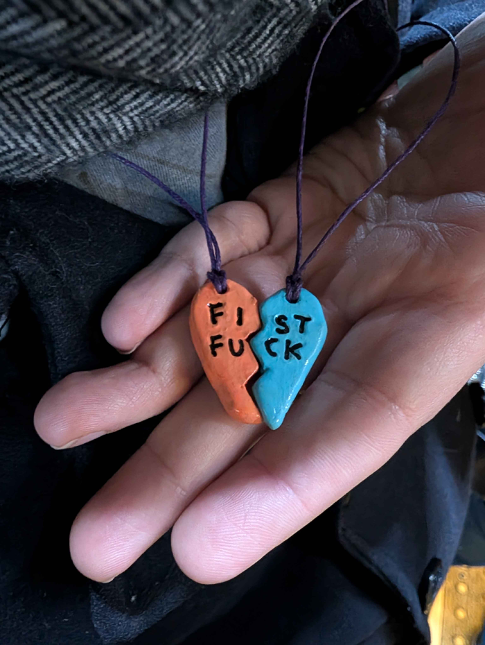 A hand holding the two pieces of a cut-in-half heart necklace, except instead of Best Friends it says FIST FUCK.