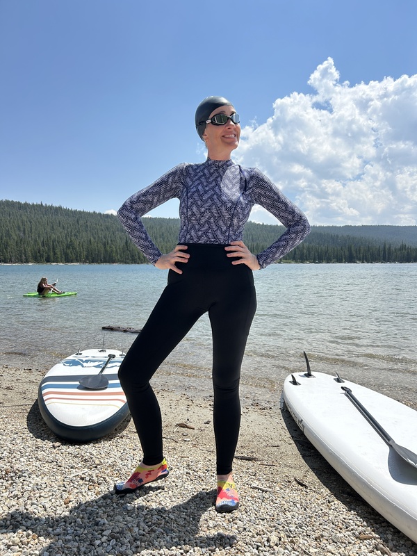 A woman standing in front of a mountain lake surrounded by paddleboards. She's wearing long swim leggings, a long sleeve swimsuit, a swim cap, and goggles.