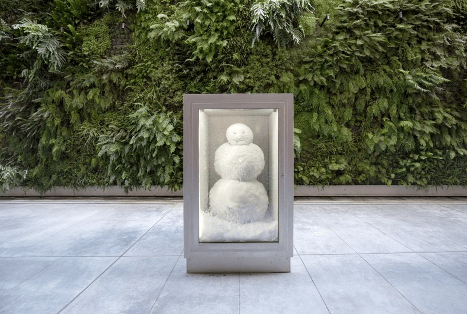 A photo of a snowman inside a metal refrigerated box, sitting outdoors on a flat area of gray pavement, with green vegetation in the background. The snowman has two large snowballs for its body and a smaller snowball for the head, with two eyes and a smiling mouth drawn on the head, but no other markings. The overall effect is incongruous; it is strange to see a snowman outdoors on what is clearly a warm summer day