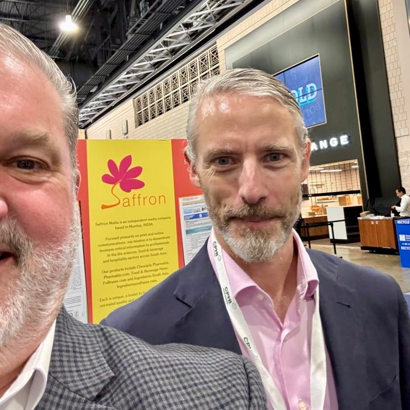 Photo of 2 white men at a trade show, standing in front of a booth for a company called Saffron. The man taking the picture has cropped it so only half his face is in the frame.
