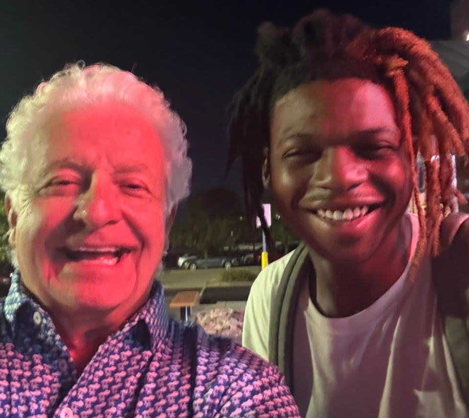 A nighttime selfie of two smiling men standing close together outdoors, lit by a warm reddish light — an older man with wavy white hair in a patterned button-up shirt on the left, and a younger man with long dreadlocks in a white t-shirt and backpack strap on the right, with a dark parking lot and a few cars visible in the background.
