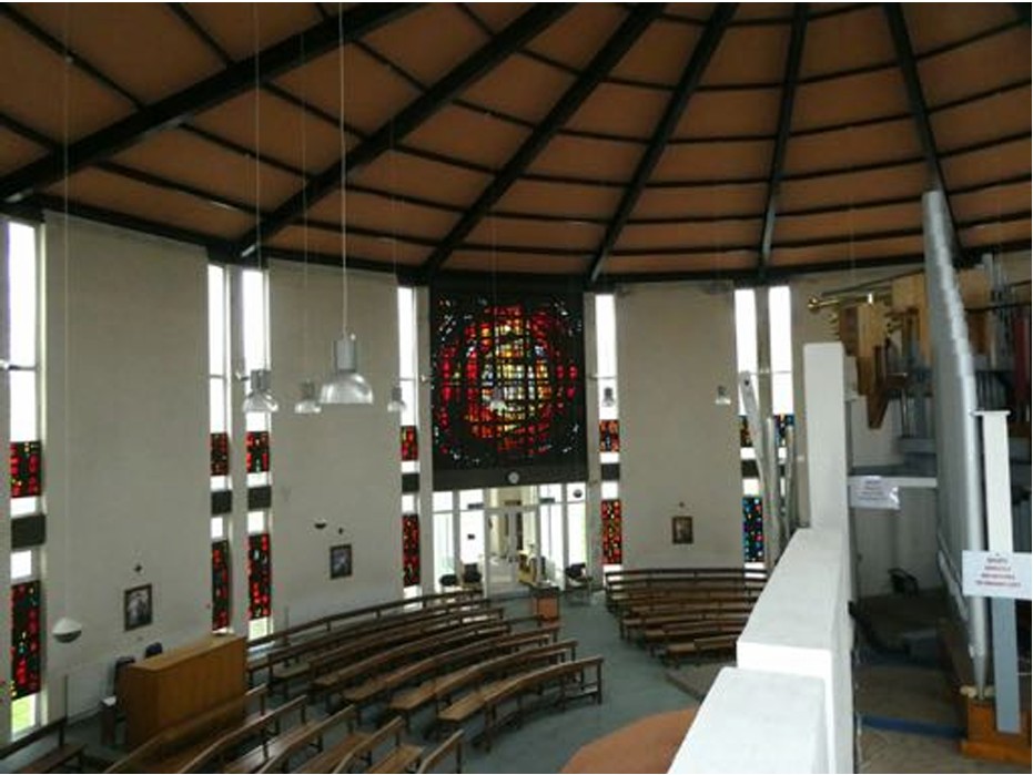 Photo of the interior from the organ loft. A large wooden ceiling rises to an apex just out of shot. The curved radial pews are visible, along with the dalle de verre set into the narrow windows. Above the main entrance, a huge double-height dalle de verre fills a whole wall. It has reds and yellows in some form of abstracted circular design.