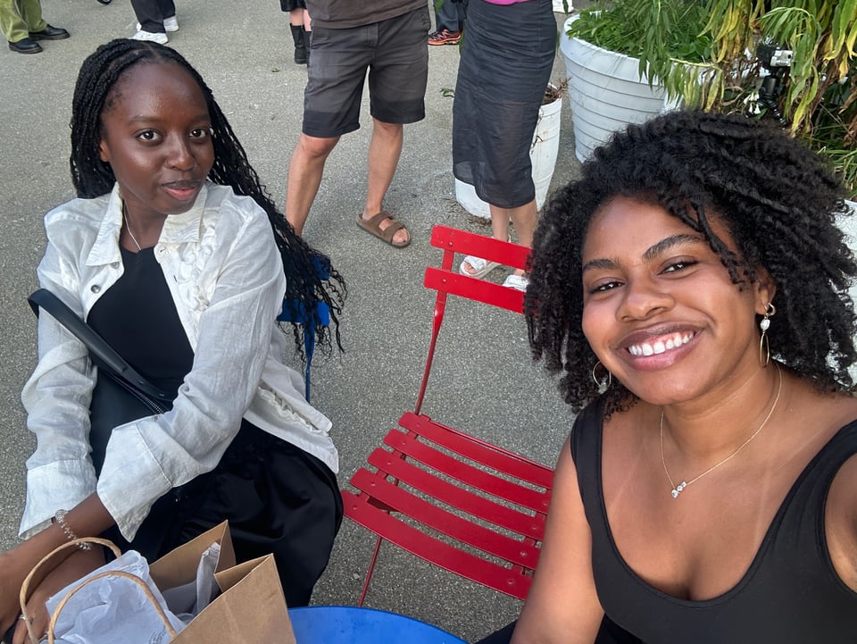 [Two people sitting next to each other in a park, a red lawn chair and shopping back between them. It’s a sunny day and they look easeful and content.]