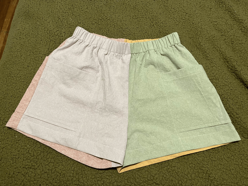 A pair of shorts sit on a textured fabric surface. The colours white, pink, green and yellow are visible. The shorts have pockets and an elastic waistband!