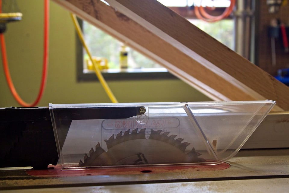 A photograph of a table saw showing a many-toothed blade protruding out of a metal base. The blade has a plastic cover on top.