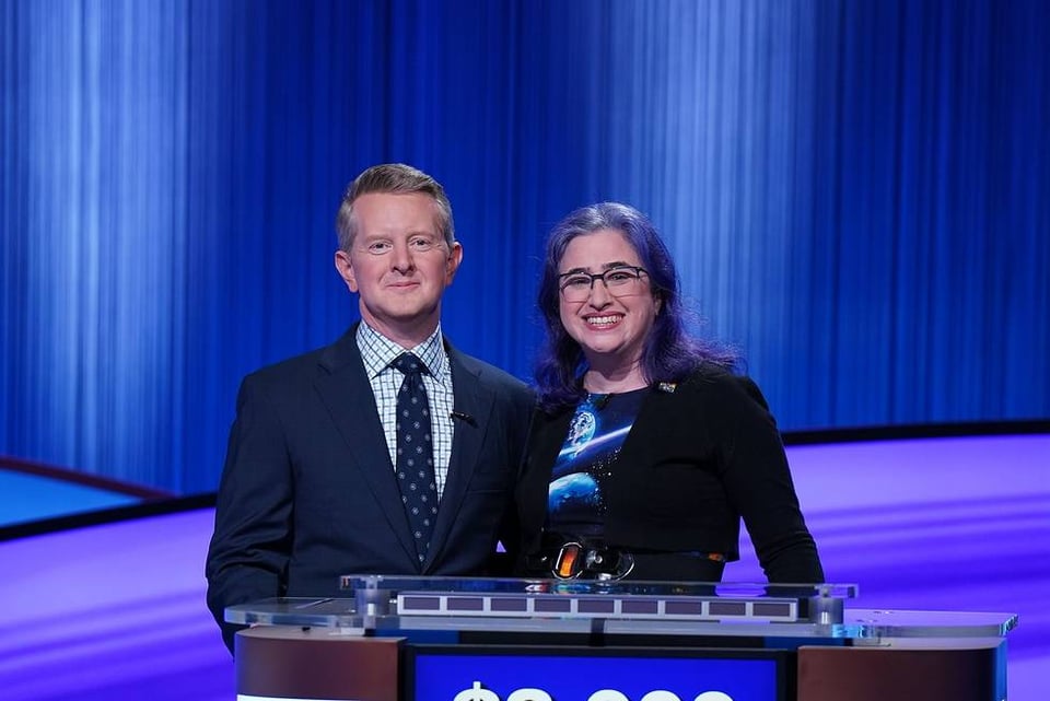 Me and Ken Jennings posed and smiling behind my podium on the Jeopardy! stage. I'm in my space-themed dress and black cardigan. Ken is wearing a blue suit, a white shirt with a blue geometric pattern on it, and a blue tie with some kind of small, repeating motif. Between the outfits, my purple hair and the blue and purple stage, it's a very blue and purple picture