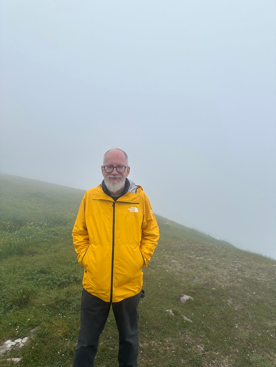 I am standing a respectful distance from the edge, the landscape lost in fog behind me. I am wearing a bright yellow rain jacket and have my hands firmly in my pocket, and I hold my body tight as I tensely smile for the camera.