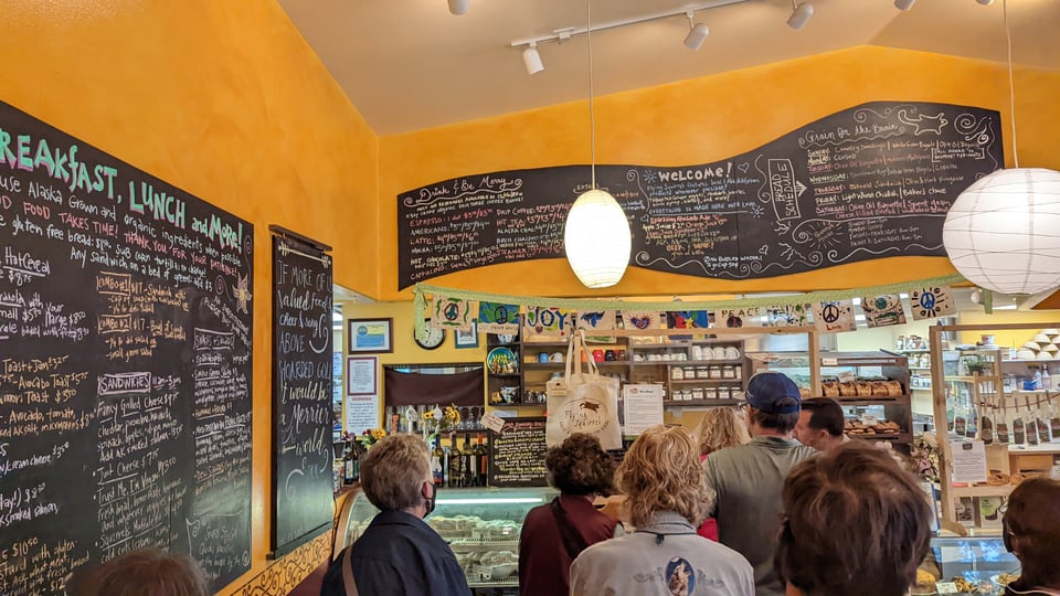 Busy, colorful signs line a cafe's yellow walls. People queue to order in front of a case. Flags and items for purchase fill the area behind the counter