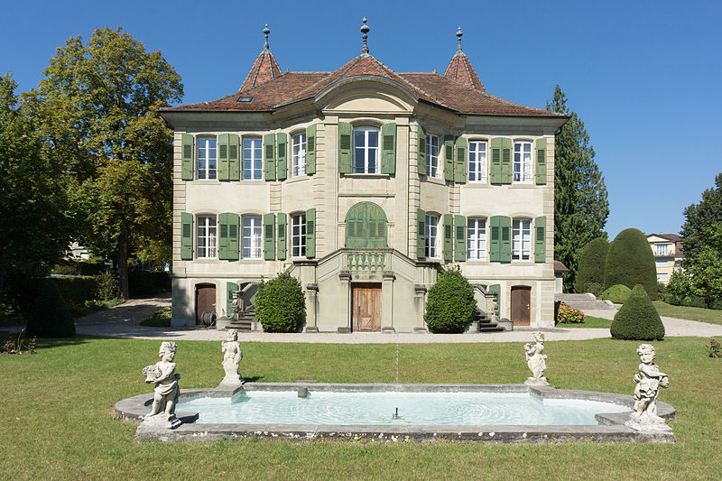 A photograph of a grand chateau, with green shutters, and a small pond ringed with four statues of cherubs
