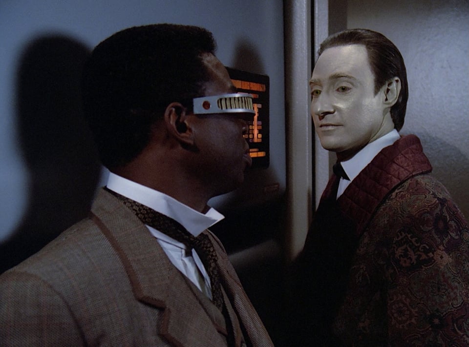 Geordi and Data, dressed as Watson and Holmes, prepare to enter the holodeck. Data has a sly smile.