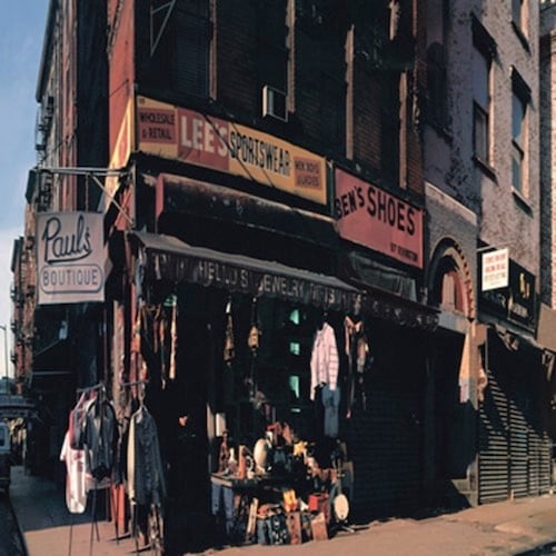A photograph of a street corner in New York. There is a store called Paul's Boutique on the corner, as indicated by the sign that hangs over the sidewalk to the left. Above the store is a tall brick edifice, presumably apartments. On the sidewalk are clothes hanging from awning as a display, as well as a table with all sorts of objects on it, but difficult to identify any one object. There are two additional signs on the building, just below the brick edifice: "Lee's Sportswear" and "Ben's Shoes."