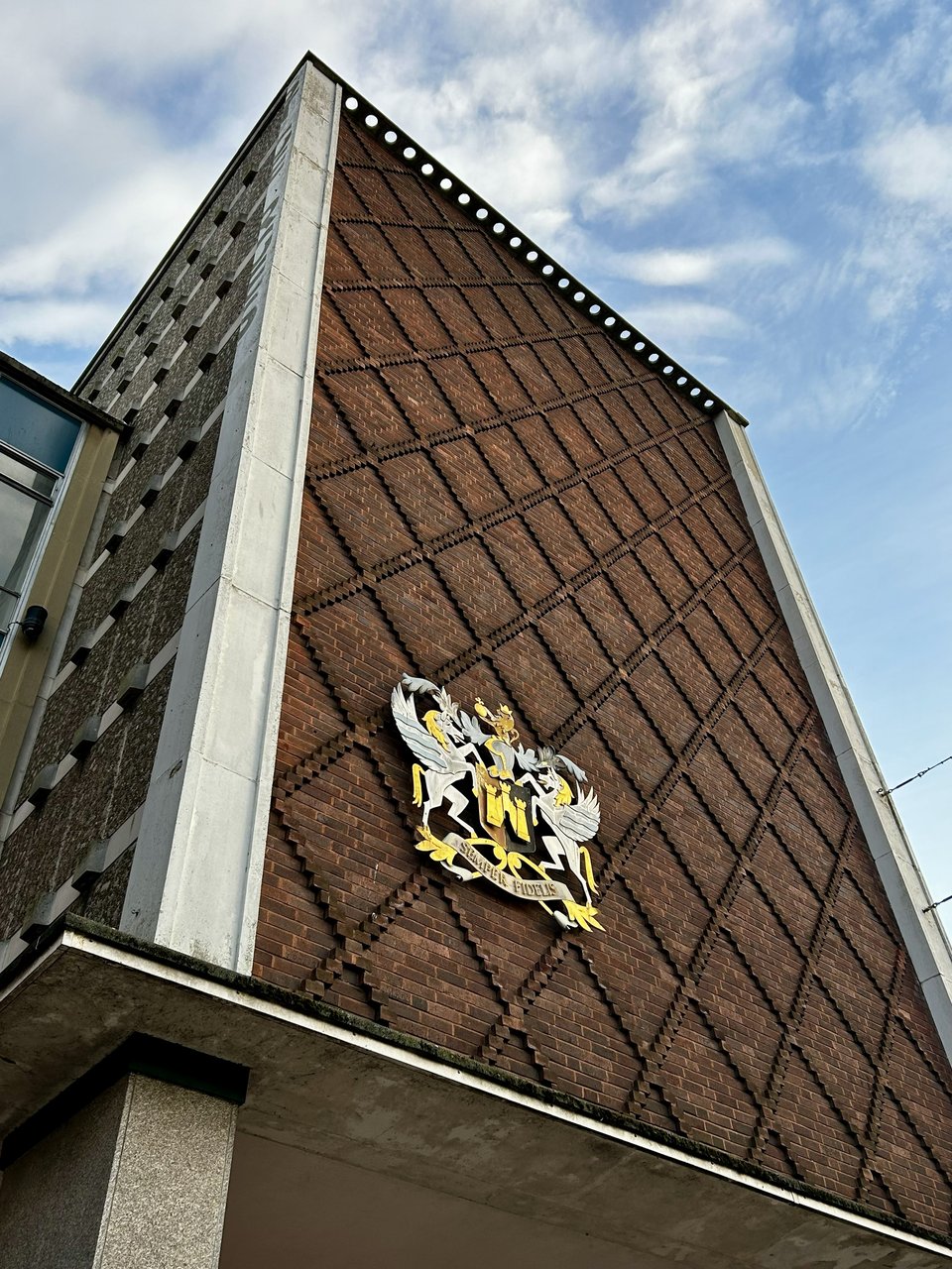 A 1960s flytower against a blue sky. The concrete frame has been filled on one side with red brickwork with a raised diamond pattern. The other wall is filled with rough concrete panels in a square grid pattern. At the very top of the concrete frame, there's a ghost sign in darker concrete, because they took the old name down after power washing the building.
