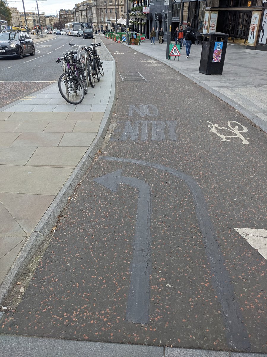 An asphalt protected cycle lane no longer features a 'no entry' marking and right-sweeping arrow direction, having been de-marked using perhaps burning or bitumen leaving the shape of the original marking in black against the black and red chip of the lane.