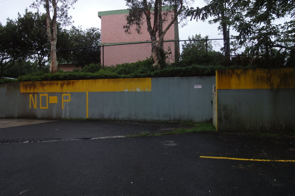 Gray wall with yellow stripe reads "NO-P" in large letters, under a cloudy sky. Trees and a pink building are behind.
