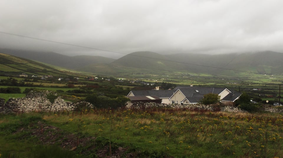 A photograph of the misty Kerry countryside. The clouds are low and grey over the rolling hills, stone walls are crumbling, and houses are dotted across the patchwork fields.