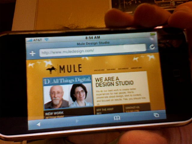 A slightly blurry photo of the first iPhone showing the golden Mule Design website with the all things d website with Walt Mossberg and Kara Swisher and the words We Are A Design Studio