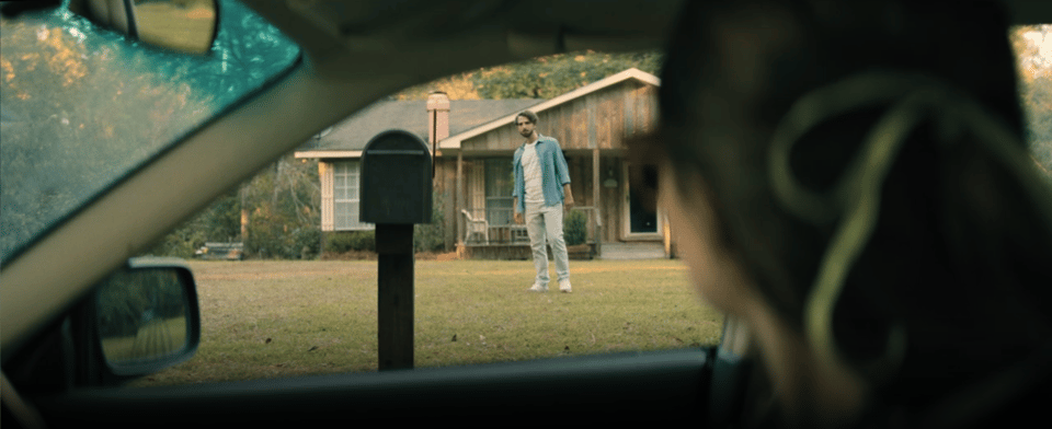 Brynn, a white woman with shoulder length brown hair pulled back by a ribbon, is about to attempt to wave at her neighbor, a white man with short brown hair and a short beard. He wears a white tee with a long sleeve blue button down over it and grey pants and white sneakers. He does not wave back.