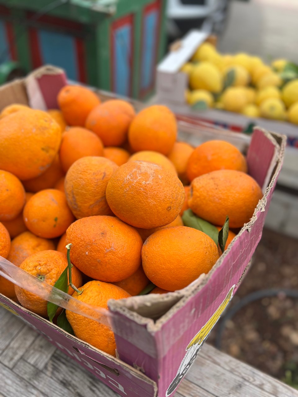 A photograph of a big box of oranges, with a box of lemons in the background