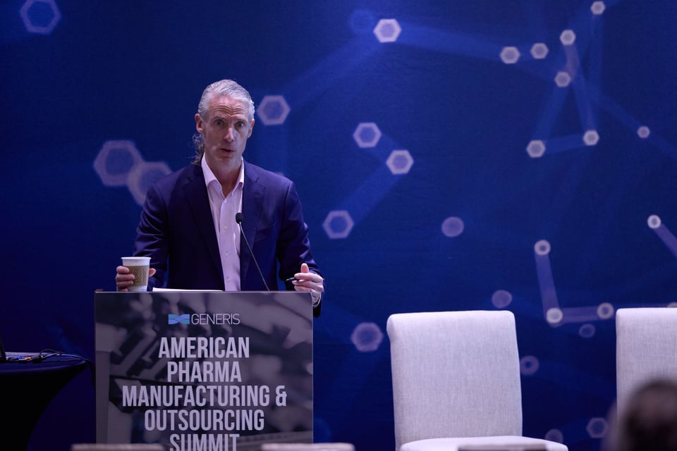 Photo of white man in navy suit & pink shirt at a lectern speaking into a microphone. He has a cup of coffee in his right hand a pen in his left