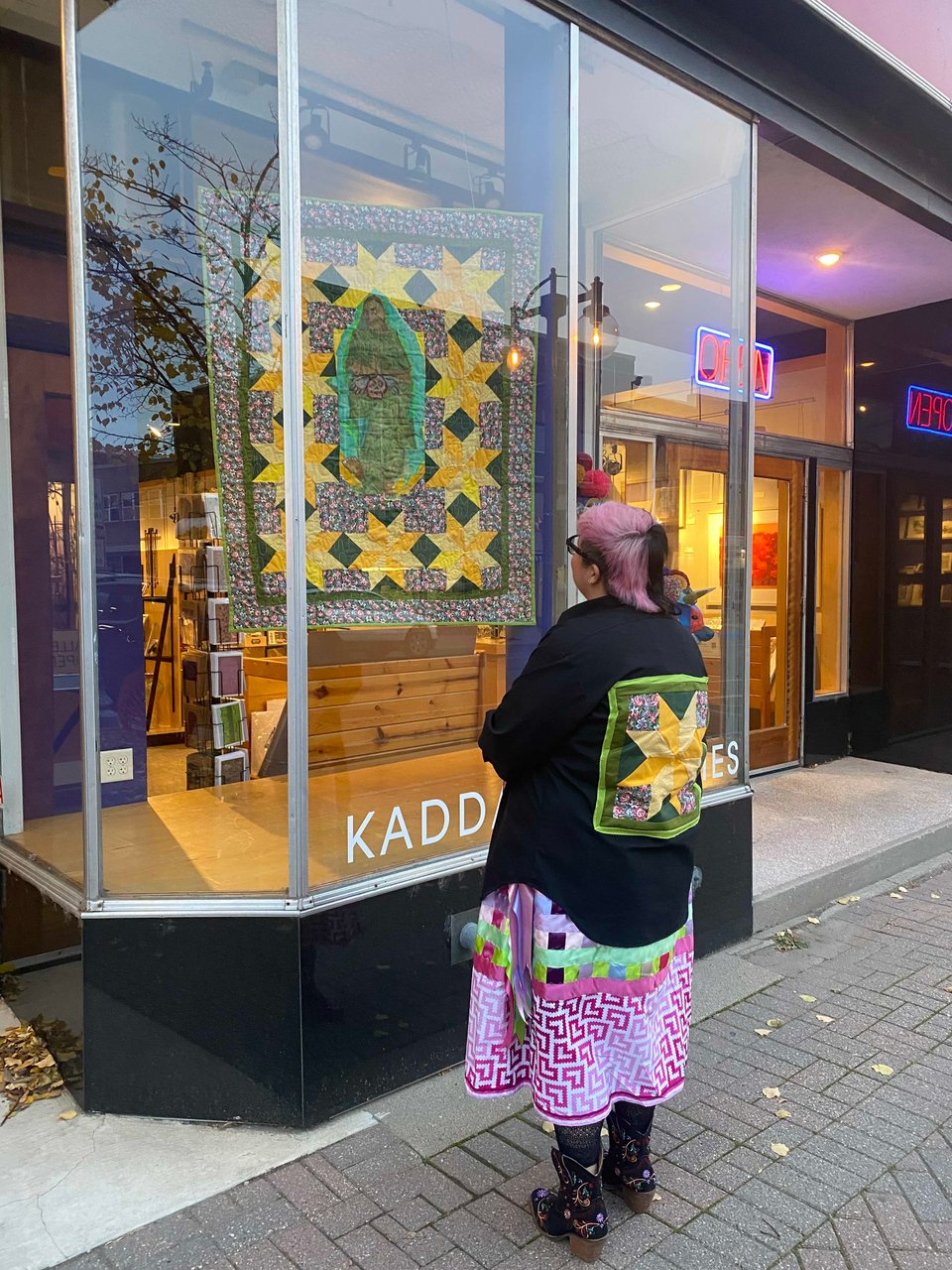 a femme artist looks at a quilt with a painted figure of la virgen de guadalupe wearing a quilt block from the work of art on their jacket