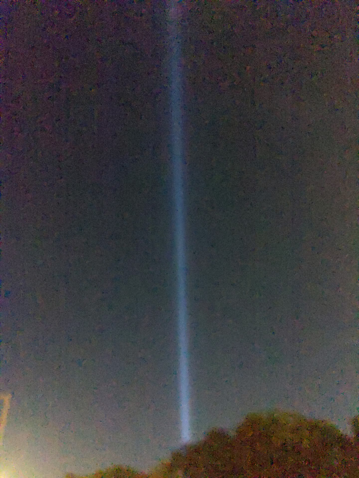 Grainy photo of the sky with one of the World Trade Center memorial lights seen off in the distance.