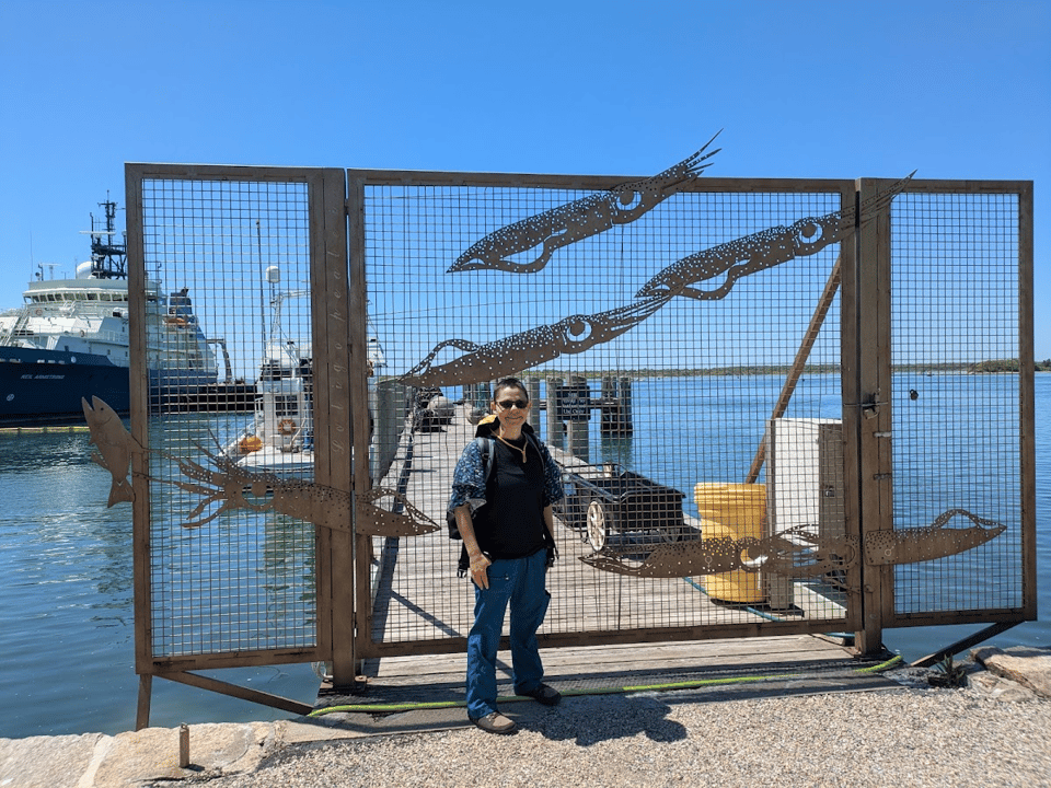 Photo of me standing in front of a metal gate at the entrance to a pier. A large research vessel is visible in the background. I am wearing a shirt with plankton on the sleeves, though it's hard to tell in the picture. The gate is decorated with large beautiful metal cutouts of market squid, including two in the lower right who are mating head-to-head and labeled with male and female indicators.