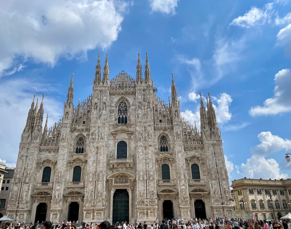Shows the facade of Duomo Di Milano, tha Cathedral of milan. There are many tourists in the front, and blue skies and clouds in the background