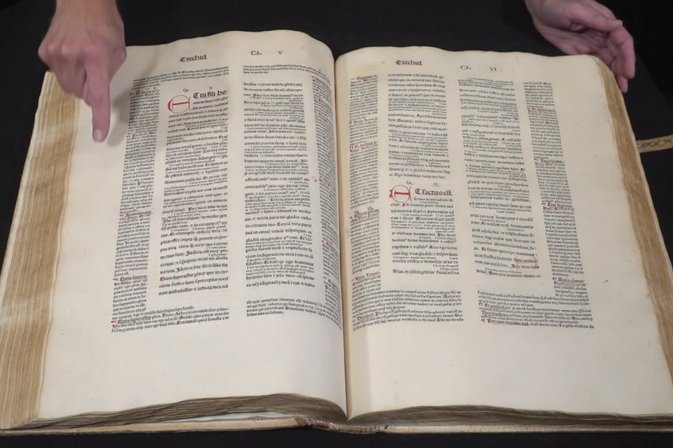 [A large book opened up to a page showing text in Latin with commentary laid out in the margins around it. A person’s finger points to the commentary.]