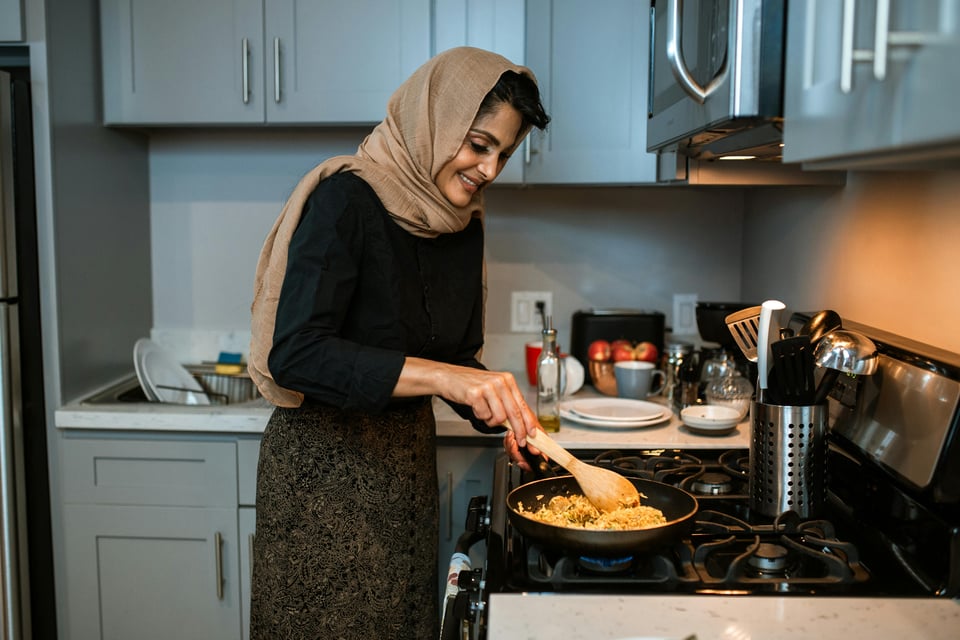 A woman wearing a headscarf stands over a stove. She's stirring a pan, cooking, and smiling as she does.