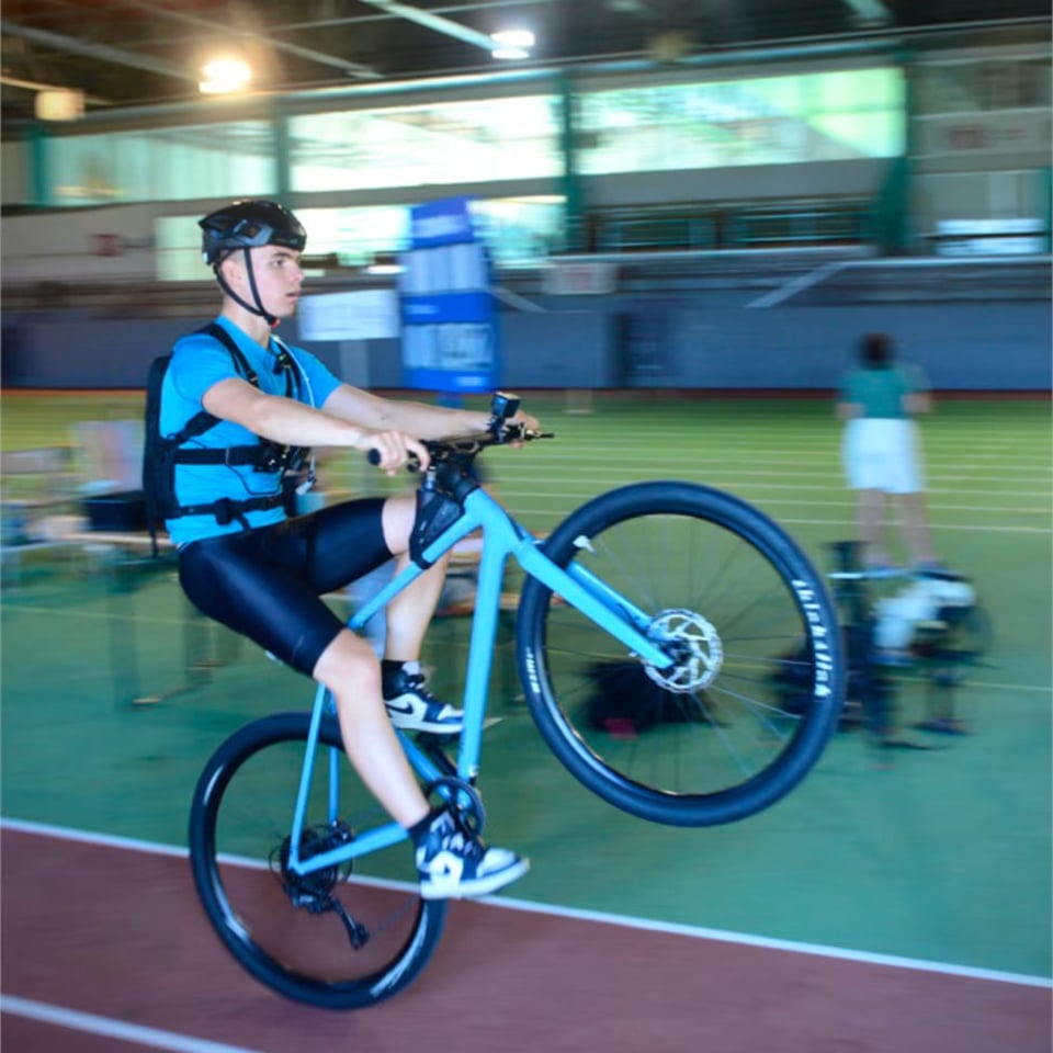 A cyclist in a blue jersey and black helmet performs a wheelie on a light-blue bike while riding indoors on a track. The background is motion-blurred, emphasizing speed, with gym equipment and a few people faintly visible. The rider looks focused and balanced, holding the handlebars steady as the front wheel lifts high off the ground.