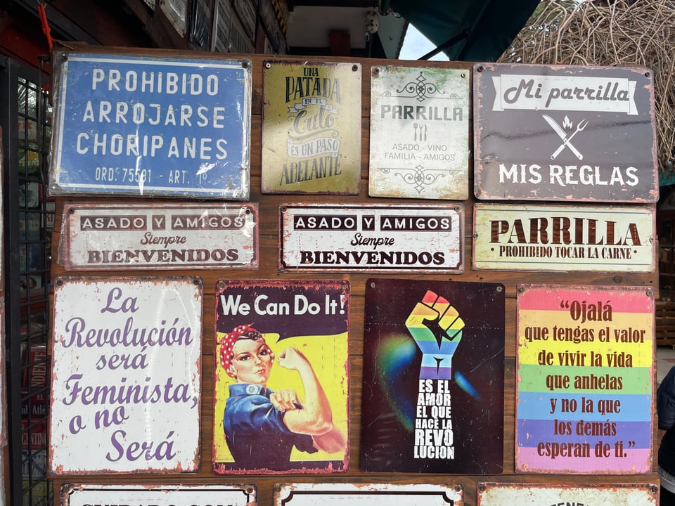 A wooden panel is filled with various aluminum plates with sayings on them "Mi parrilla, mis reglas" (My grill, my rules)