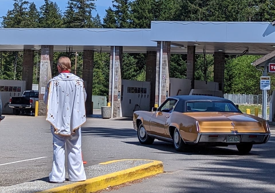 Chuck Gay as Elvis in the parking lot of the car wash. Back to the camera, white Elvis jumpsuit and cape, gold caddy driving towards the carwash.