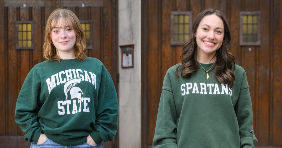 Two young women stand side by side, each wearing a green Michigan State sweatshirt with different logos. They are positioned outdoors in front of a wooden door with small windows, both smiling at the camera.