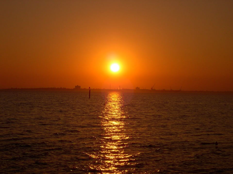 The sun sets over the ocean. The sky is burnt orange. The sun is bright in the middle and the sun's reflection forms a line in the water. The silhouette of some buildings and Melbourne Port can be seen in the distance.