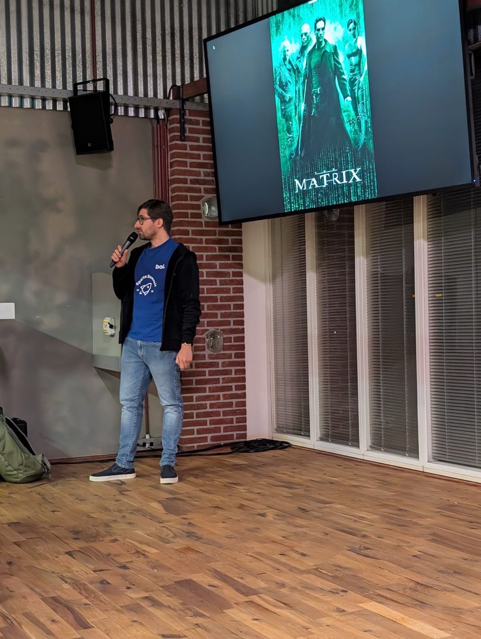 A man stands in front of a room holding a microphone. Above and behind him the screen shows a still from the movie The Matrix.