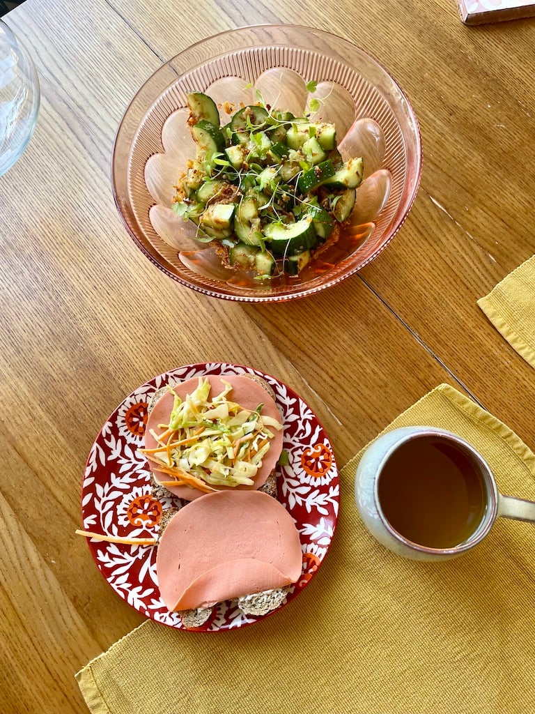 A cucumber salad with two slices of ham on bread, some coleslaw and a cup of green tea