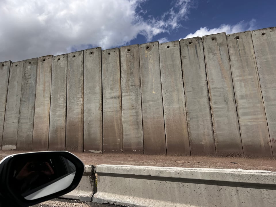 A wall made of tall concrete slabs along the side of a road. The car's side mirror is in the picture, and the sky is blue with clouds