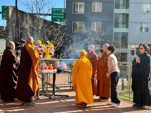 Five monastics wearing a mixture of brown, orange, and burgundy, robes stand with palms together facing a two-tiered altar on an elevated outdoor deck. Four monastics have shaved heads and the monastic in orange robes in the center is wearing an orange knit cap. In the background is a modern building with multiple windows to the right and a brown freeway barrier to the left. Above the barrier in the distance is a green freeway sign with the text “90 West Masspike Worcester.” The two-tiered altared rests on two separate tables and contains pink and yellow flowers, golden candles, various fruit offerings, and a black incense burner with smoke coming out of it. One of the two monastics on the left, in side profile, is holding a bell striker. Two of the monastics on the right, in side profile, are holding microphones. A layperson in a beige sweater, black pants, and white tennis shoes leans forward toward the altar. Another layperson in black robes and glasses, with palms pressed together, stands to the right. A videographer behind the altar is filming the scene. All eight people pictured are of Asian heritage.