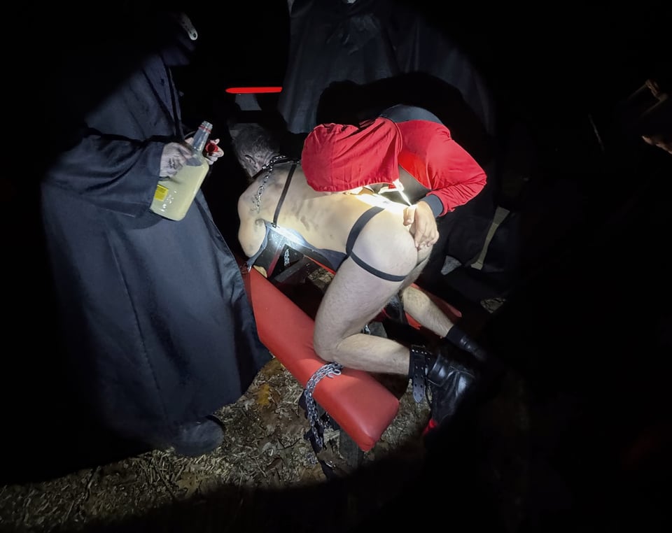 a man in jockstrap kneels on a red leather bench as another man takes a shot from his back