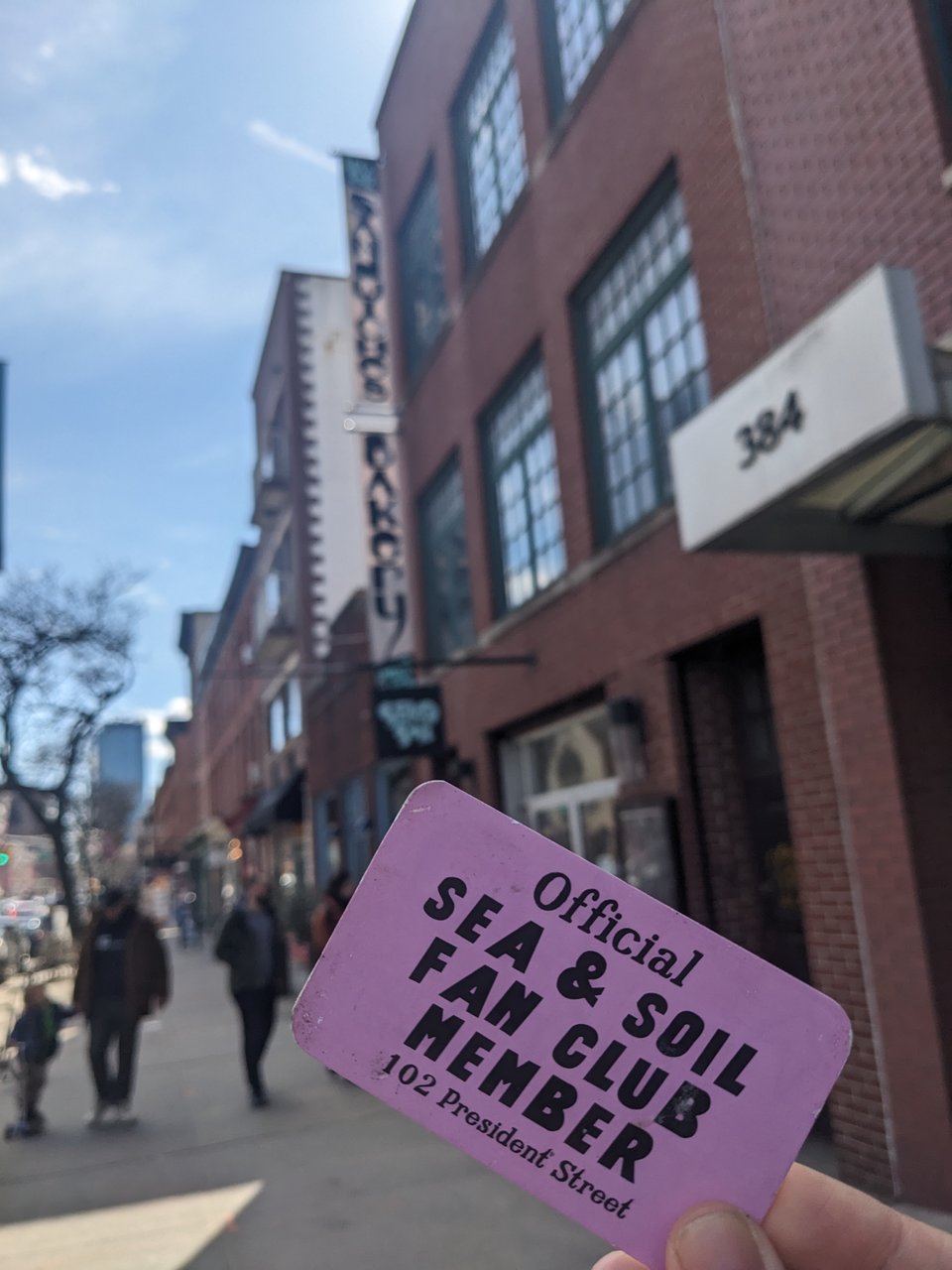 My right hand holding a pink "Sea & Soil Fan Club Member" card in front of the new Sea & Soil restaurant, inside a brick building with a vertical white banner