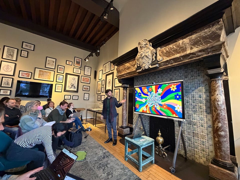 A man stands in front of crowd of people in a room. There is a large screen in the front and it says "Vibe Coding Collective" with a wavy colorful pattern from the 1960s behind the words.