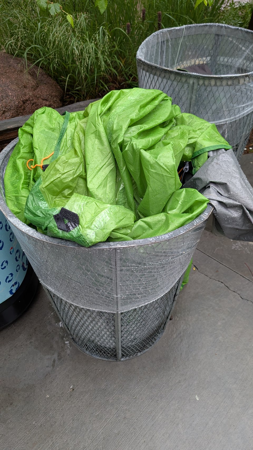 A shot of a trash bin with a green tarp or tent stuffed in it. The material appears wet.