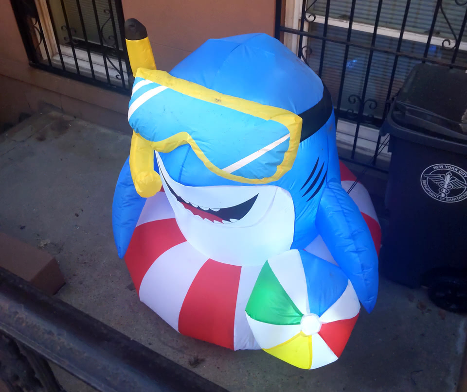 A blue inflatable smiling shark with a snorkel mask, inner-tube, and beach ball in front of a residence in Brooklyn